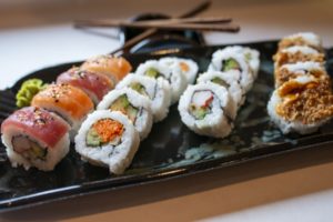 a horizontally framed photograph shows a close and low angle view of a clay coyote large tray glazed in midnight garden that is being used to serve sushi. there are 17 pieces of sushi and a dab of wasabi on the tray. the tray is laying horizontally across the photograph with both small sides and handles cut out of frame. the sushi row to the very left of the photograph is nigiri style with the fish draped over the rice, the other 3 rows are regular roll style. the row of 4 pieces of sushi on the very right of the photograph have one side of each piece (the side towards the viewer) coated in a brown crunchy layer. the clay coyote little dipper is near the top center of the photograph, resting behind the large tray. it has two pairs of chopsticks sitting on the square little dipper making an X. the photograph is well lit with white light. the tray and little dipper are resting on a white kitchen counter top.