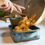 a vertically framed photograph shows a clay coyote small skillet that has been used to start the apple slices for an apple crisp. the sliced and peeled apples are being poured out of the skillet into a clay coyote square baker. behind the square baker with the apples being actively poured into it is a clay coyote whisky bowl glazed in joes blue. it is partially obscured by the baking dish and apples. the bakers hands are visible in the upper left corner, they are holding the small skillet aloft with one hand and using a wooden spatula with the other. the workspace is a white kitchen counter top.