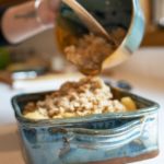 a vertically framed photograph shows a clay coyote whiskey bowl being used to pour a crunchy topping onto an apple crisp contained in a clay coyote square baking dish. both the whiskey bowl and the baking dish are glazed in joes blue. the bakers hands are visible coming from the upper left of the photograph with only their forearms and hands visible. the baker is using their left hand to hold the whisky bowl aloft over the baking dish, and is using their right hand to scoop the crunchy topping from the whisky bowl into the square baking dish. the baking dish is sitting on a white kitchen countertop, and is well lit by natural light.
