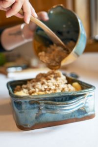 a vertically framed photograph shows a clay coyote whiskey bowl being used to pour a crunchy topping onto an apple crisp contained in a clay coyote square baking dish. both the whiskey bowl and the baking dish are glazed in joes blue. the bakers hands are visible coming from the upper left of the photograph with only their forearms and hands visible. the baker is using their left hand to hold the whisky bowl aloft over the baking dish, and is using their right hand to scoop the crunchy topping from the whisky bowl into the square baking dish. the baking dish is sitting on a white kitchen countertop, and is well lit by natural light.