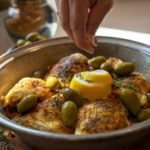 a horizontally framed photograph shows a close up picture of a clay coyote tagine bottom with 6 pieces of well spiced chicken in it, with whole green olives, lemon zest and fresh herbs. the hand of the chef is coming into the frame from the top center, and has a full hand pinch grip, and is about to drop another ingredient onto the top center of the dish. the tagine bottom is glazed in coyote grey, which denotes it is flameware. behind the main focus of the photograph (the tagine dish) and to the slight left is a out of focus clay coyote large vase glazed in joes blue. there is sunlight visible on the white kitchen counter top behind the tagine bottom.