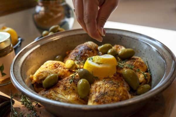 a horizontally framed photograph shows a close up picture of a clay coyote tagine bottom with 6 pieces of well spiced chicken in it, with whole green olives, lemon zest and fresh herbs. the hand of the chef is coming into the frame from the top center, and has a full hand pinch grip, and is about to drop another ingredient onto the top center of the dish. the tagine bottom is glazed in coyote grey, which denotes it is flameware. behind the main focus of the photograph (the tagine dish) and to the slight left is a out of focus clay coyote large vase glazed in joes blue. there is sunlight visible on the white kitchen counter top behind the tagine bottom.