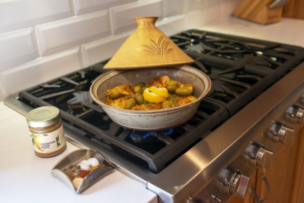 the photograph shows a clay coyote tagine glazed in yellow salt, with the lid resting behind the base, on its back lip. the tagine is on a gas stovetop range. directly next to the stove top oven range, on the white kitchen countertop, is a jar of New York Shuk's preserved lemon paste (sold at clay coyote and on the clay coyote website). in front of the jar of NYS preserved lemon paste is a clay coyote little dipper glazed in mint chip. the little dipper is the square style. it is filled with various spices. the background is a white brick façade backsplash.