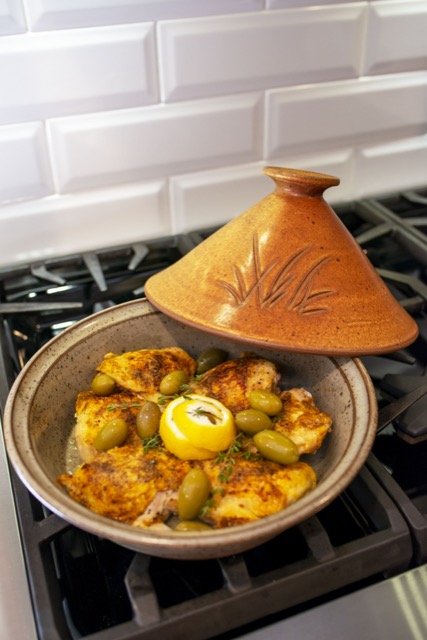 the photograph shows a clay coyote tagine glazed in moroccan sand resting on a gas stove top range. the lid of the tagine is resting on the back lip of the base. the base is filled with chicken, olives, lemon and fresh herbs. the background is a white brick façade.