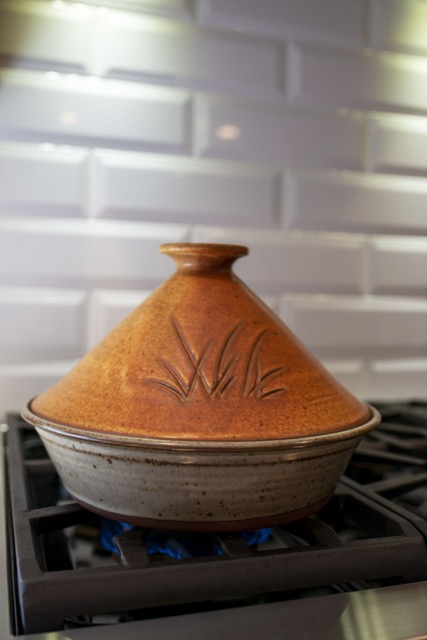 a vertically framed photograph shows a clay coyote tagine with the lid glazed in Moroccan sand. the bottom of the tagine is glazed in coyote grey, which denotes it is flameware. the entire tagine is sitting on a kitchen stove top range, with a barely visible low flame underneath it. behind the tagine is a white brick motif back splash.