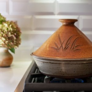 a horizontally framed photograph shows a clay coyote tagine glazed in Moroccan sand. the tagine is sitting on a stovetop kitchen range, with the lid resting directly onto the base. behind and to the left of the tagine is a clay coyote small vase glazed in feather, with a bouquet of flowers resting in it. the burner that the tagine is resting on is set to low, with just barely any blue flame visible. the backsplash to the kitchen range is a white brick motif. the front of the tagine lid has a grass motif.