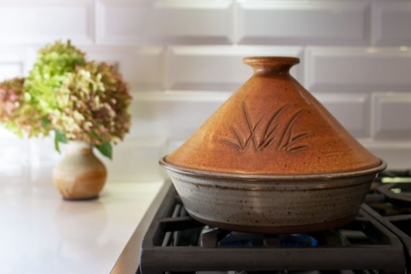 a horizontally framed photograph shows a clay coyote tagine glazed in Moroccan sand. the tagine is sitting on a stovetop kitchen range, with the lid resting directly onto the base. behind and to the left of the tagine is a clay coyote small vase glazed in feather, with a bouquet of flowers resting in it. the burner that the tagine is resting on is set to low, with just barely any blue flame visible. the backsplash to the kitchen range is a white brick motif. the front of the tagine lid has a grass motif.