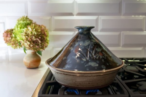 a horizontally framed photograph shows a clay coyote tagine with the lid glazed in midnight garden (black with pops of red and blue) . the bottom of the tagine, and all clay coyote tagines is glazed in coyote grey (light grey with dark speckling through out it), to denote that they are flameware. the tagine is sitting on a lit gas burner, with small blue flame visible underneath the tagine. behind and to the left is a small clay coyote vase with a large chrysanthemum bouquet resting in it. the vase is sitting on a smooth white kitchen countertop. the backsplash for the oven range is a white brick motif.