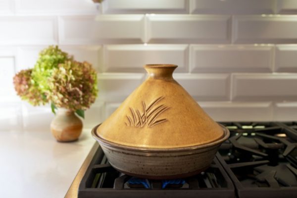 a horizontally framed photograph shows a clay coyote tagine with lid glazed in Moroccan sand resting on an unlit kitchen gas range. behind and to the left (sitting on the white kitchen countertops) is a small clay coyote vase with a large bouquet of flowers in it. the backsplash is a white brick motif.
