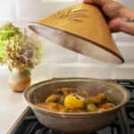 the photograph shows a clay coyote tagine resting on a stove top gas range. the lid is glazed in yellow salt. in the base of the tagine there is chicken and olives with fresh herbs and lemon slices. there is visible condensation on the part of the lid visible, and small wisps of steam are visible coming off the chicken. behind and to the left of the tagine base, on the nearby white kitchen countertop, is a clay coyote small vase glazed in feather. it has a large bouquet of flowers resting in it. the flowers have red and green colors. the background is a white brick facade