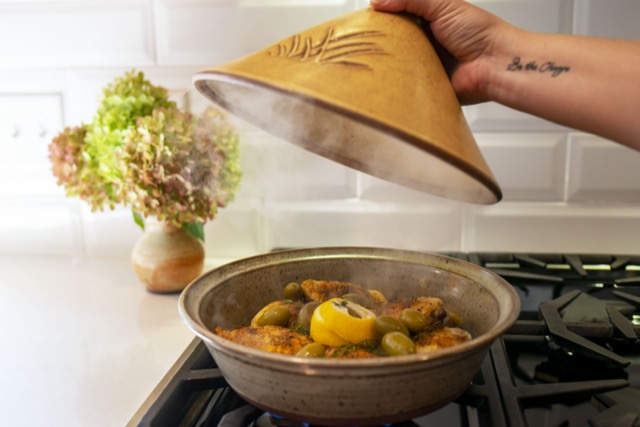 the photograph shows a clay coyote tagine resting on a stove top gas range. the lid is glazed in yellow salt. in the base of the tagine there is chicken and olives with fresh herbs and lemon slices. there is visible condensation on the part of the lid visible, and small wisps of steam are visible coming off the chicken. behind and to the left of the tagine base, on the nearby white kitchen countertop, is a clay coyote small vase glazed in feather. it has a large bouquet of flowers resting in it. the flowers have red and green colors. the background is a white brick facade