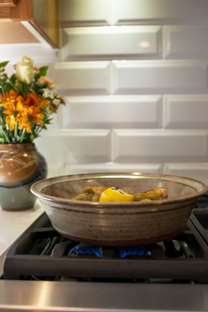 a vertically framed photograph shows a clay coyote tagine bottom being used to cook chicken on a gas stove top range. the chicken has spices, whole green olives and sliced fresh lemon flavoring it. under the tagine bottom a small amount of blue flame can be seen coming from the burner. behind and to the left of the tagine, sitting on a white kitchen countertop is a clay coyote large vase glazed in joes blue. the backsplash for the kitchen range is a white brick motif.
