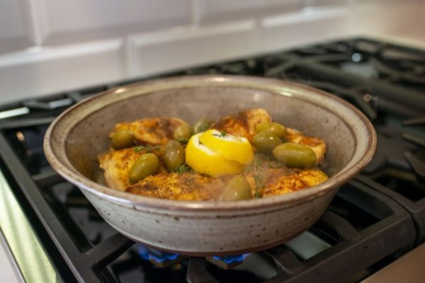 a horizontally framed photograph shows a close up of the base of a tagine being used on a gas range stovetop. inside of the tagine bottom there is chicken thighs cooking. they have been well spiced and have fresh lemon, green olives and fresh herbs on them. a blue flame from the gas burner is visible underneath the tagine bottom. the tagine bottom is made out of clay coyote flameware and is glazed in coyote grey (a light grey with dark speckles through out).