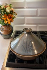 a vertically framed photograph shows a clay coyote tagine glazed in Moroccan blue sitting on a unlit gas range. behind it and to the left is a large vase glazed in joes blue, which has a bouquet of flowers resting in it.