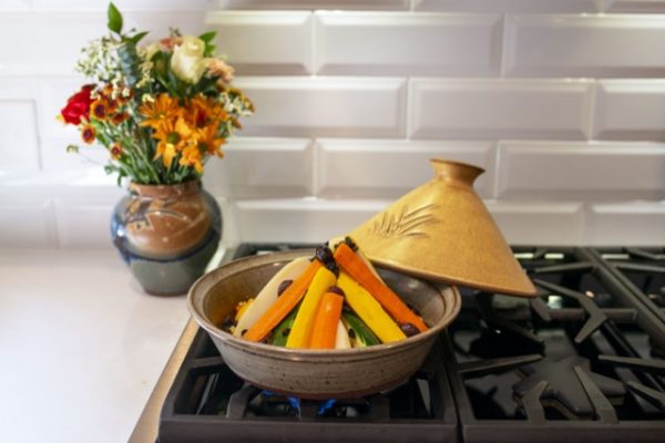 a horizontally framed photograph shows a clay coyote tagine glazed in yellow salt sitting on a gas kitchen stove top range. the tagines lid (the yellow salt) is resting on the back "quarter" of the tagine bottom, (relative to the viewer) is filled with vegetables stacked ready to cook. the tagine bottom is glazed in coyote grey, which denotes it is flameware. behind and to the left of the tagine, (sitting on a white kitchen counter top) is a clay coyote large vase glazed in joes blue, with a large bouquet of flowers. the backsplash is a a white brick motif.