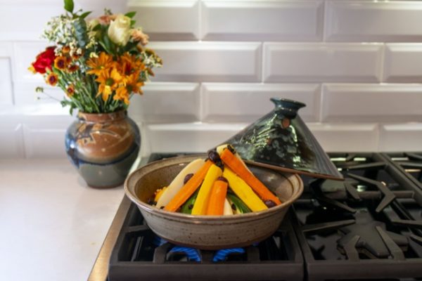 a horizontally framed photograph shows a clay coyote tagine with top glazed in midnight garden, resting on a kitchen stove top gas range. the midnight garden lid has been taken of from the bottom (glazed in coyote grey to denote it is flameware) where there is a conical shaped pile of vegetables cooking inside. the lid is partially resting on the "back" lip of the tagine bottom and the unused burners. behind the tagine base to the left on the white kitchen counter top is a clay coyote large vase glazed in joes blue, with a bouquet of flowers placed in it. the veggies in the tagine bottom are all cut to be long and thin, and are arranged in a semi standing conical shape, where the tips are resting against each other and they angle down to the base. behind the gas range stove top is a white brick motif backsplash. blue gas flame is visible underneath the tagine.