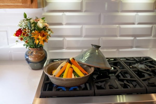 a horizontally framed photograph shows a clay coyote tagine with top glazed in Mediterranean blue, resting on a kitchen stove top gas range. the blue lid has been taken of from the bottom (glazed in coyote grey to denote it is flameware) where there is a conical shaped pile of vegetables cooking inside. the lid is partially resting on the "back" lip of the tagine bottom and the unused burners. behind the tagine base to the left on the white kitchen counter top is a clay coyote large vase glazed in joes blue, with a bouquet of flowers placed in it. the veggies in the tagine bottom are all cut to be long and thin, and are arranged in a semi standing conical shape, where the tips are resting against each other and they angle down to the base. behind the gas range stove top is a white brick motif backsplash. blue gas flame is visible underneath the tagine.