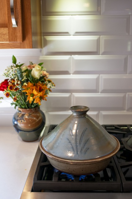 a vertically framed photograph shows clay coyote tagine with a Moroccan blue glazed top. the tagine bottom like all clay coyote bottoms are glazed in coyote grey, as they are flameware. behind and the the left of the of the tagine is a clay coyote large vase glazed in joes blue. the vase is sitting on a white counter top which is directly next to (connected) the gas kitchen range the tagine is sitting on. there is blue flame visible underneath the tagine.