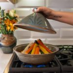 a horizontally framed photograph shows a clay coyote tagine with the top glazed in moroccan blue in the process of cooking vegetables. the tagine top (the moroccan blue part) is being held aloft above the tagine bottom, and above the vegetables. morgans right hand is coming from out of frame to the right and has just lifted the tagine top up to reveal the cooking vegetables contained within. behind and to the left of the tagine, is a clay coyote vase glazed in joes blue, with a bouquet of flowers resting in it. the flowers have red, orange, white, yellow and green. the backsplash behind the oven range is a white brick motif