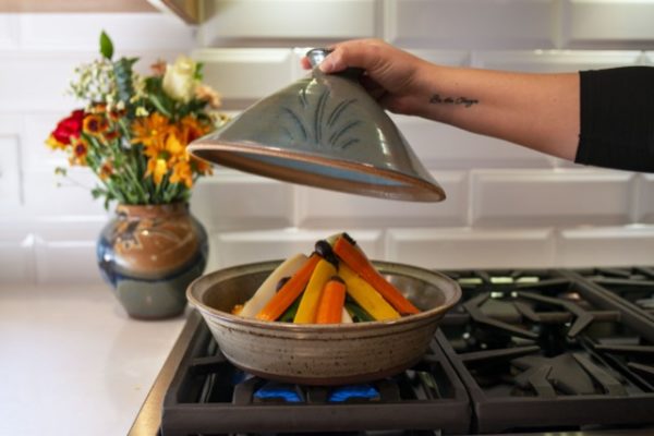 a horizontally framed photograph shows a clay coyote tagine with the top glazed in moroccan blue in the process of cooking vegetables. the tagine top (the moroccan blue part) is being held aloft above the tagine bottom, and above the vegetables. morgans right hand is coming from out of frame to the right and has just lifted the tagine top up to reveal the cooking vegetables contained within. behind and to the left of the tagine, is a clay coyote vase glazed in joes blue, with a bouquet of flowers resting in it. the flowers have red, orange, white, yellow and green. the backsplash behind the oven range is a white brick motif