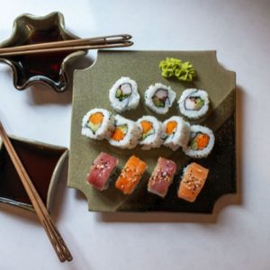 a horizontally framed photograph shows a clay coyote sushi plate being used to serve sushi. to the left of the sushi plate are two clay coyote little dippers. each little dipper has a pair of chopsticks resting across them, and have soy sauce poured into the bottoms to be used as dipping bowls. the sushi plate has 12 pieces of sushi on it, 4 of which are nigiri style. those 4 nigiri style are closest to the view. at the "top" of the sushi plate is a double dab of wasabi. the sushi tray and both little dippers are all glazed in mint chip. everything is resting on a white kitchen counter top. the photograph is lit with bright white light mostly concentrated on the upper left of the photograph, but the entire photograph is well lit.