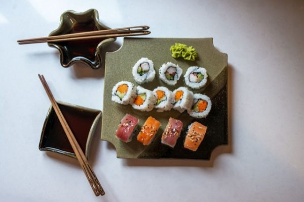 a horizontally framed photograph shows a clay coyote sushi plate being used to serve sushi. to the left of the sushi plate are two clay coyote little dippers. each little dipper has a pair of chopsticks resting across them, and have soy sauce poured into the bottoms to be used as dipping bowls. the sushi plate has 12 pieces of sushi on it, 4 of which are nigiri style. those 4 nigiri style are closest to the view. at the "top" of the sushi plate is a double dab of wasabi. the sushi tray and both little dippers are all glazed in mint chip. everything is resting on a white kitchen counter top. the photograph is lit with bright white light mostly concentrated on the upper left of the photograph, but the entire photograph is well lit.
