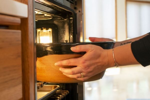 a horizontally framed photograph shows a pair of hands placing a clay coyote special edition cassole into a open oven. there is a metal colored baking sheet on the rack below the one the cassole is being placed into. the forearms and hands of the person placing the cassole are the only things visible from them. they have a wedding ring and silver colored bracelet on, and the ends of their sleeves are visible, and are black.