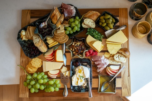 a horizontally framed photograph shows a overhead top down view of a charcuterie spread. everything is on a wooden butchers block, which in turn is sitting on a white kitchen countertop. the spread is using clay coyote serving pottery, including little dippers, a large tray with handles, and a small tray without. there is slice baguettes hard and soft cheeses, green grapes, green olives, honey, mixed nuts, multiple kinds of crackers and some thinly sliced deli meat.