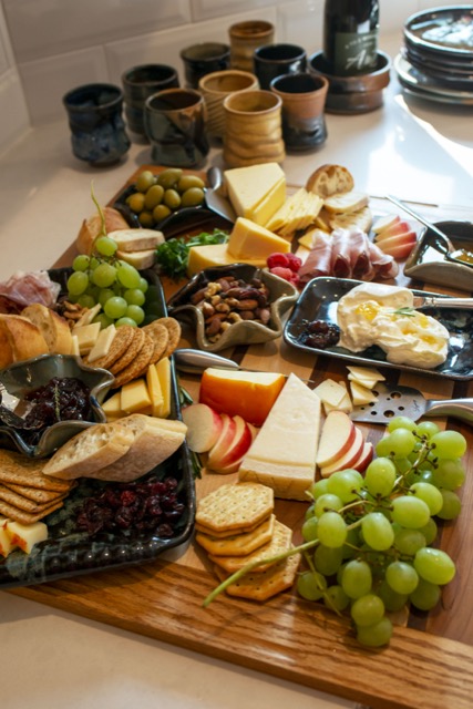 a eye level photograph shows clay coyote pottery being used in a charcuterie spread. the entire layout is resting on a cutting board. the charcuterie spread includes: hard and soft cheeses, green grapes, green olives, sliced apples, sliced baguettes, wheat and water crackers, mixed nuts, sliced meat and raspberries. the clay coyote pottery used are: large tray glazed in midnight garden, small tray in midnight garden, and multiple little dippers being used as condiment holders and a mixed nut mix holder.