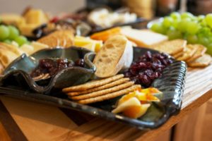 a close up of clay coyote serving ware being used as part of charcuterie spread. horizontally framed. the large tray is glazed in midnight garden, and has handles built in. the large tray is holding: craizens, sliced apples, wheat crackers, sliced baguettes and a clay coyote little dipper, glazed in zappa. behind the large tray is the rest of the charcuterie spread (slightly out of focus) all of this is resting on a wooden cutting board