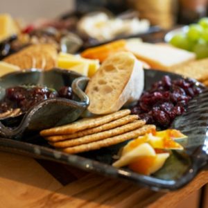 a close up of clay coyote serving ware being used as part of charcuterie spread. horizontally framed. the large tray is glazed in midnight garden, and has handles built in. the large tray is holding: craizens, sliced apples, wheat crackers, sliced baguettes and a clay coyote little dipper, glazed in zappa. behind the large tray is the rest of the charcuterie spread (slightly out of focus) all of this is resting on a wooden cutting board