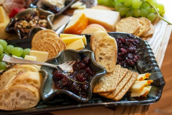 a horizontally framed photograph shows a relatively close shot of clay coyote pottery being used to serve a charcuterie spread. the large tray has craizens, sliced baguettes, wheat crackers, sliced apples, cheese, water crackers, green grapes and a clay coyote little dipper filled with a jam condiment, with a silver serving spoon resting inside. behind the large tray there are more crackers, cheese, nut mix, green grapes