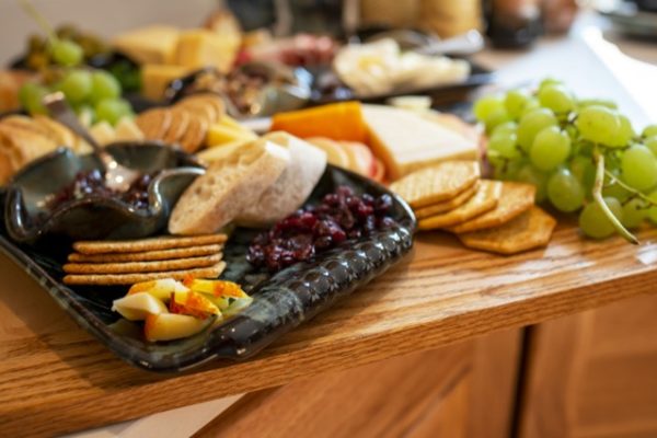 a close up horizontally framed photograph show clay coyote serving dishes being used as part of a charcuterie spread. the main focus of the photograph is a large clay coyote tray glazed in midnight garden. it is holding craizens, sliced baguettes, wheat crackers, slice apples and a little dipper. the little dipper resting on the large tray has a jam or jelly with a silver serving spoon. on the cutting board (which everything is resting on) to the right of the large tray is cheese, cracker and green grapes. in the background, further down the counter top (out of focus) is more charcuterie spread, including grapes, cheese, sliced meat and crackers.