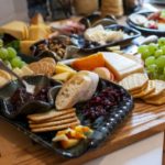 a horizontally framed photograph shows a charcuterie spread sitting on a white kitchen countertop. there is a large butchers block holding most of the spread. there is a clay coyote large tray with handles glazed in midnight garden (black base with pops of dark blue) filled with the cheese, fruit and cracker spread. these include: craisens,, sliced apples, sliced cheese, wheat crackers, water crackers, green grapes, baguette slices and a clay coyote little dipper in the center of the tray filled with a deep purple jam. there is a silver serving spoon resting in the little dipper. there are more crackers, grapes, apples, cheese and bread behind the large tray. three is a little dipper in mint chip (black and mint green) filled with a nut mix (including whole almonds and cashews). further back is a clay coyote small tray without handles also glazed in midnight garden, it is filled with a baked brie wheel and some dark purple jam in a corner. even further into the background is a clay coyote dinner plate filled with some of the charcuterie selection. a couple yonomis are visible out of focus and partially out of frame.