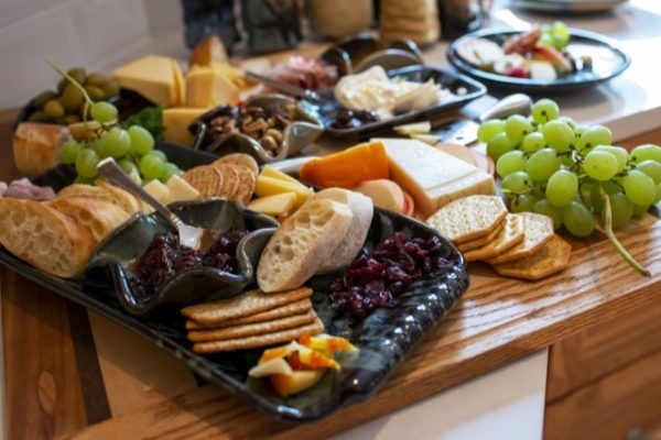 a horizontally framed photograph shows a charcuterie spread sitting on a white kitchen countertop. there is a large butchers block holding most of the spread. there is a clay coyote large tray with handles glazed in midnight garden (black base with pops of dark blue) filled with the cheese, fruit and cracker spread. these include: craisens,, sliced apples, sliced cheese, wheat crackers, water crackers, green grapes, baguette slices and a clay coyote little dipper in the center of the tray filled with a deep purple jam. there is a silver serving spoon resting in the little dipper. there are more crackers, grapes, apples, cheese and bread behind the large tray. three is a little dipper in mint chip (black and mint green) filled with a nut mix (including whole almonds and cashews). further back is a clay coyote small tray without handles also glazed in midnight garden, it is filled with a baked brie wheel and some dark purple jam in a corner. even further into the background is a clay coyote dinner plate filled with some of the charcuterie selection. a couple yonomis are visible out of focus and partially out of frame.