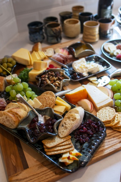 a vertically framed photograph shows a white kitchen counter filled with a charcuterie spread. there is a clay coyote large tray with handles in midnight garden (black base with dark blues, reds and yellows) filled with green grapes, water crackers, baguette slices, craisens, apple slices, cheese slices and wheat crackers. there is also a clay coyote little dipper on the large tray, it is filled with a deep purple jam, with a silver serving spoon resting in it. behind the large tray is more grapes, apple slices, cheese, silver cheese knife, green olives. there is a clay coyote little dipper in the center (in mint chip glaze- mint green and black) filled with a nut mix. there is a clay coyote small tray without handles glazed in midnight garden with baked brie and jam. further back there is another clay coyote little dipper in mint chip. all of this is resting on a butchers block which is sitting on the white kitchen countertop. sitting directly on the white countertop is 8 clay coyote yonomis and a bottle of wine (sitting in a clay coyote wine coaster) is partially out of frame.