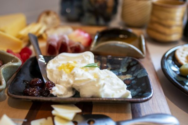 a horizontally framed photograph shows a close up of a section of a charcuterie spread. the main focus of the photograph is a clay coyote small tray (without handles) glazed in midnight garden (black base color with pops of yellow red and dark blues) the tray has baked brie placed in the middle with a silver butter knife resting on the far left (from the view) corner. in the far right corner of the small tray is a scoop of dark purple jam. there is a small sprig of a fresh herb placed on the center of the baked brie. in the (out of focus) background there is a clay coyote little dipper glazed in mocha swirl (browns and black mixing), a couple yunomis, sliced apples, raspberries and a hard cheese with slices of it next to it. in the foreground a small cheese knife with small slices of swiss is partially visible. everything listed but the yonomis are sitting on a butchers block cutting board. the yonomis are sitting on the white kitchen counter.