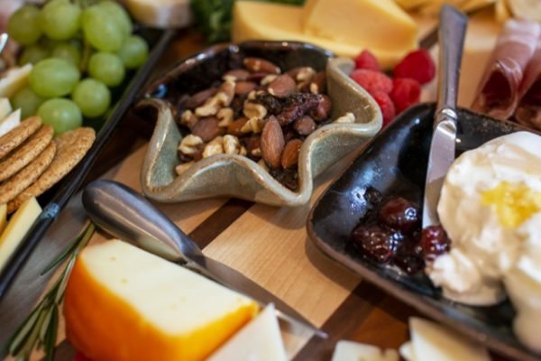 a horizontally framed photograph shows a close up of a charcuterie spread, with a clay coyote little dipper in mint chip glaze ( mint green with black) and a clay coyote small tray without handles in midnight garden. The little dipper is filled with a nut mix, and the small tray is filled with brie cheese and in the corner of the tray, a jelly. there is various hard cheeses, green grapes, wheat crackers, raspberries and a cheese knife and butter knife all visible.