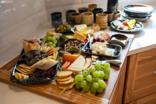 a horizontally framed photograph shows a charcuterie spread on a white kitchen counter. there is a clay coyote large tray with handles in midnight garden, various glazed little dippers, small trays, yunomis, clay coyote plates in various glazes and a clay coyote wine coaster. the charcuterie spread has green grapes, green olives, sliced apples various hard cheeses, brie cheese with jam, slices of bread, various crackers and nuts. behind this spread is a group of clay coyote yunomis in various glazes. to the right of the yunomis is a bottle of wine, sitting in a wine coaster, further right from that is a stack of clay coyote dinner plates. all of this is on a white kitchen counter top, and there is bright outside light streaming in from an unseen near by window