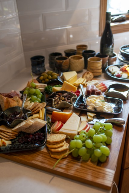 a vertically framed photograph shows a charcuterie spread on a white kitchen counter. there are clay coyote yunomis, wine coasters, small trays, a large tray with handles (midnight garden glazed) clay coyote plates and little dippers. the food of the charcuterie spread are grapes, cheeses, sliced apples, brie cheese, jams, green olives, and various shapes of crackers. behind the charcuterie spread there is various shaped and glazed clay coyote yunomis, and a bottle of wine in a clay coyote wine coaster. slightly out of frame to the right of the wine and cups are some clay coyote plates. there is daylight coming in from a window in the upper right corner.