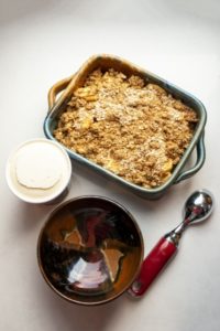 a vertically framed photograph shows a clay coyote baking dish in joes blue filled with apple crisp. it is a top down view. lower in the photograph is a clay coyote chili bowl in merlot glaze ( red, brown and black). it is empty. to the right of the bowl is a ice cream scoop with a metal head and red serving handle. in between the baking dish and the bowl, slightly to the left is a full pint of vanilla ice cream. it has a smooth surface, with no scoops missing. all of this is sitting on a white kitchen counter top.
