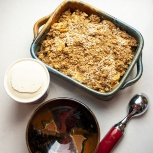 a vertically framed photograph shows a clay coyote baking dish in joes blue filled with apple crisp. it is a top down view. lower in the photograph is a clay coyote chili bowl in merlot glaze ( red, brown and black). it is empty. to the right of the bowl is a ice cream scoop with a metal head and red serving handle. in between the baking dish and the bowl, slightly to the left is a full pint of vanilla ice cream. it has a smooth surface, with no scoops missing. all of this is sitting on a white kitchen counter top.