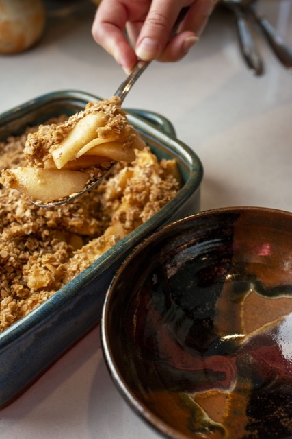 a vertically framed photograph shows a clay coyote square baking dish in joes blue glaze filled with freshly baked apple crisp, a hand is in the middle top of frame, scooping out a serving of it. to the right and slightly lower in frame to the baking dish is a clay coyote chili bowl in merlot glaze (black, browns and red). both the dishes are sitting on a white kitchen counter top.