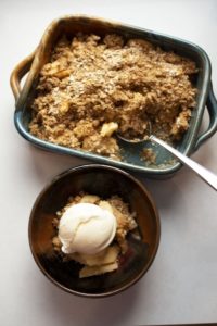 a vertically framed photograph shows a top down view of a clay coyote chili bowl in merlot and a clay coyote baking dish glazed in joes blue. the baking dish is filled with freshly baked apple crisp, with a scoop missing out of the lower right corner. a large silver serving spoon rests there, tucked into the apple crisp. the chili bowl is filled with the missing scoop of apple crisp and a single spherical scoop of ice cream. the bowl is towards the "bottom" of the photograph while the baking dish is towards the "top". the photograph is bathed in white