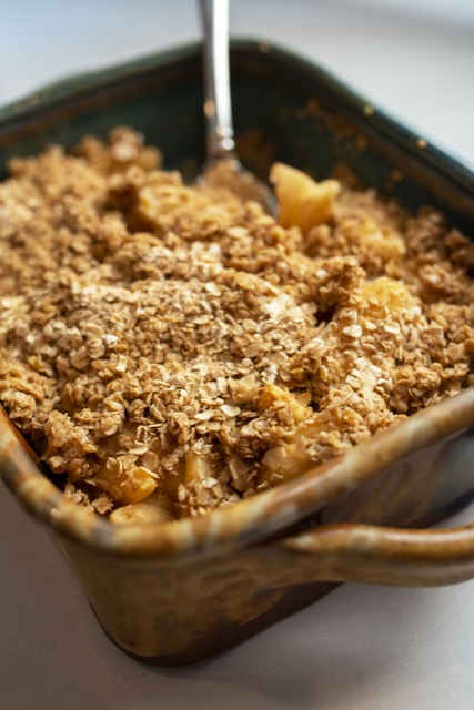 a vertical framed photograph shows a close up picture of a clay coyote baking dish in joes blue glaze. the brown section of the glaze is covering the corner and handle nearest to the viewer. the freshly baked apple crisp has a silver serving spoon in the back corner of the dish, with some of the apple crisp already served. the apple crisp has a oatmeal and cinnamon crunchy topping coving most of the sliced apples. the baking dish is sitting diagonal compared to the photographs framing. the baking dish is sitting on a white kitchen countertop.