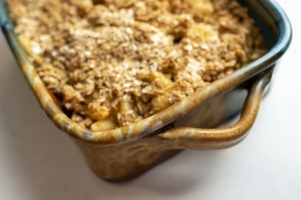 a horizontal orientated photograph of a clay coyote square baking dish. the baking dish is glazed in joes blue with the brown section of the glaze in the nearest corner to the viewer. the baking dish is filled with baked apple crisp, with a crunchy oat/cinnamon topping. the baking dish is photographed from a very close high angle, with only half of the dish visible in the photograph. the baking dish is at a 45 degree angle to the framing of the photograph. the square baking dish is sitting on a white kitchen countertop.