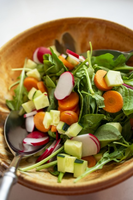 a vertically framed photograph shows a close up high angle picture of a clay coyote deep salad bowl in feather glaze, filled with a undressed spinach salad. the salad includes cut up cucumbers, carrot coins and radish slices. in the lower left of the photograph a large silver colored serving spoon is resting in on the side of the salad. the deep salad bowl in feather shows a light to cream colored base covered in a swirl of rich browns and rust color, with the cream base showing through. the left and right side of the deep salad bowl are out of frame. the deep salad bowl is sitting on a white kitchen counter top