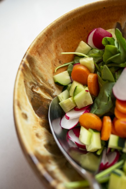 a vertical orientated photograph. a close up photograph from a high angle shows around 1/3rd of a clay coyote deep salad bowl, filled with an undressed spinach salad. the spinach salad has sliced radishes, carrot coins and diced cucumber mixed through out. the visible edge of the deep salad bowl shows off the color swirl of the feather glaze. the glaze has deep browns swirling and mixing with rusty and cream colors. between the undressed spinach salad and the deep salad bowl a silver serving spoon has been inserted, with only the large spoon head visible, while the start of the handle goes quickly out of frame back towards the viewer. the deep salad bowl is sitting on a white kitchen countertop.