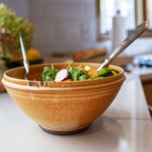a vertical framed photograph of a clay coyote deep salad bowl in feather glaze. the deep bowl is filled with a spinach salad, with cut radishes and carrots visible. the salad is undressed, with two silver salad serving utensils on either side (left and right to the viewer). the bowl is sitting on a white kitchen countertop, with the rest of the photograph out of focus. the salad bowl is reddish brown or rusty colored with just a hint of dark brown coming over the lip of the bowl coming over in one small spot slightly left of center, on the rim of the bowl.