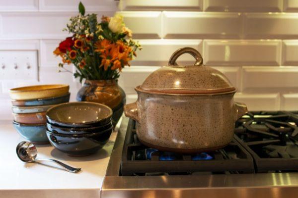 a horizontal orientated photograph. a photograph of a clay coyote dutch oven in coyote grey (grey with dark speckling through out) with the lid on, sitting on a lit gas kitchen burner. next to the dutch oven to the immediate left, on the white kitchen countertop, sits two stacks of clay coyote chili bowls. the closest stack of bowls is 3 high with the top bowl being glazed in mint chip. the bottom two bowls are in a dark glaze. behind the stack of three bowls and to the left is a stack of 5 bowls. they are glazed top to bottom in: yellow salt, joes blue, mint chip, joes blue and the bottom bowl is a dark glaze (hard to id). infront of the two stacks of bowls, also sitting on the white kitchen countertop is a silver soup ladle. Behind and between the dutch oven and the stack of three bowls is a large clay coyote vase glazed in joes blue. the vase has a fall themed bouquet placed in it, with yellow, orange and greens. behind the oven a ceramic backsplash resembling bricks is seen. barely visible underneath the dutch oven, a small faint blue flame can be seen.